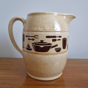 May include: A beige ceramic jug with a brown design of kitchen utensils, including a pot, a fork, a knife, a spatula, a glass, a citrus juicer, a mug, and a bowl.