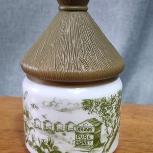 May include: A vintage honey jar with a white base and a brown, thatched-roof lid. The jar features a green illustration of a rural scene with bees and the text "SHAWS PURE HONEY".