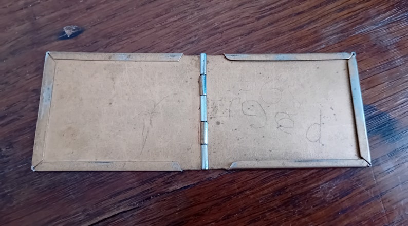 May include: A small, brown, rectangular, hinged photo album with a silver metal frame. The album is closed and has the word "Fred" written on the right side.
