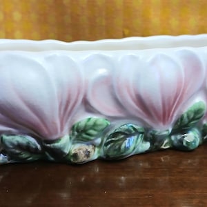 May include: A ceramic planter in the shape of magnolia flowers, predominantly white with pink and purple accents. The rectangular planter features a scalloped edge and green leaves at the base. Measurements are in centimetres.