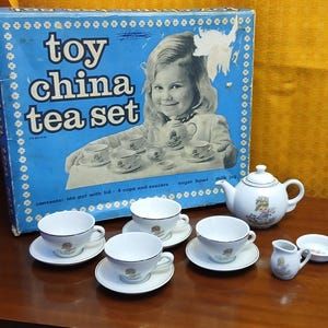 May include: A vintage toy china tea set in a blue box. The set includes a teapot, cups, saucers, a sugar bowl, and a milk jug. The tea set is white with gold trim and a floral design. The box reads 'toy china tea set'.
