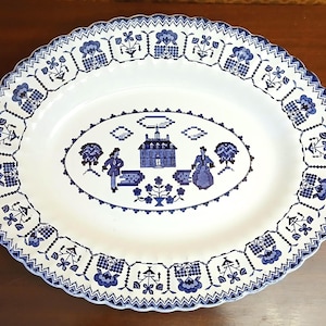 May include: An oval ceramic platter featuring a white base with a blue transferware pattern. The center depicts a house, two figures, and trees. The scalloped rim is decorated with a repeating geometric and floral motif in blue and white.