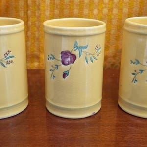 May include: Three yellow ceramic containers with a floral design. Each cylindrical container features a purple flower with blue and green leaves. The containers have a light green band at the base and rim.