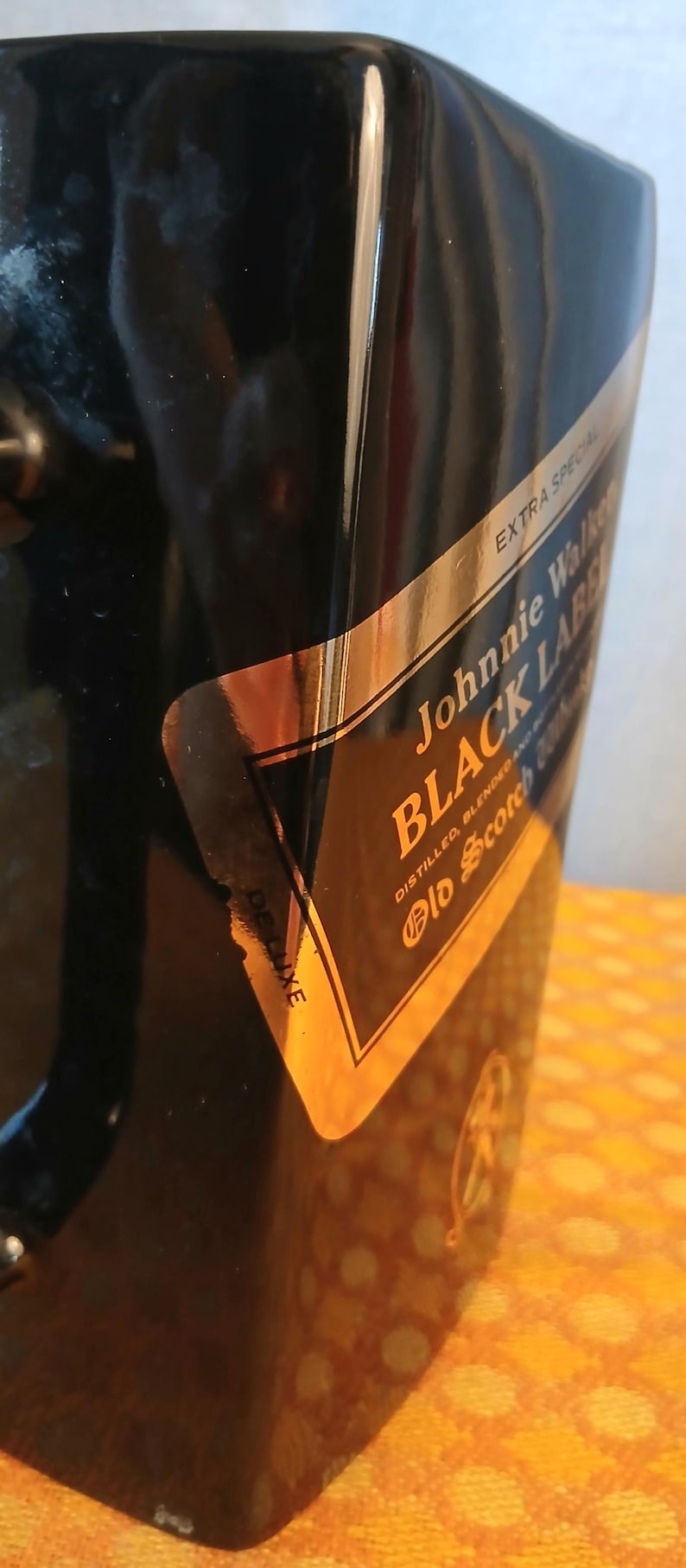 May include: Black ice bucket with a gold and black label that reads "Johnnie Walker BLACK LABEL". The label also says "Extra Special" and "Deluxe". The bucket is on a yellow surface.