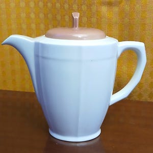 May include: White ceramic teapot with a light brown lid and handle. The teapot has a classic design with a spout, handle, and a lid with a small knob. The teapot is likely used for serving tea.