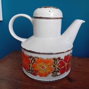Peut inclure: Théière en céramique blanche avec un motif floral dans les tons orange, rouge et marron. La théière a un couvercle avec un petit trou au sommet.