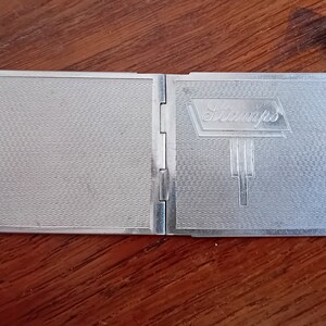 May include: A silver compact with a textured surface and the word "Stamps" embossed on the front.