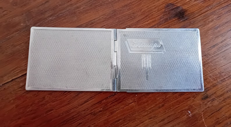 May include: A silver compact with a textured surface and the word "Stamps" embossed on the front.