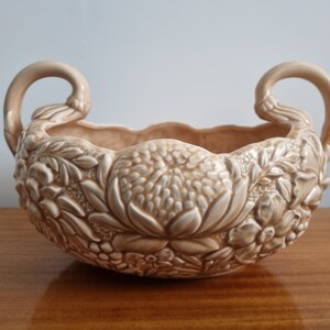 May include: A ceramic planter with two handles, featuring a raised floral design in shades of beige and white.