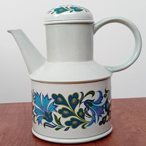 May include: A white ceramic teapot with a floral design in shades of blue, green, and purple. The teapot has a lid and a handle.