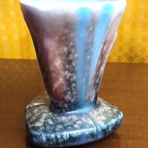 May include: A vintage ceramic vase with a flared top and a unique, textured base. The vase features a color palette of blue, brown, and white, with a speckled pattern on the base. The rim has a scalloped edge.