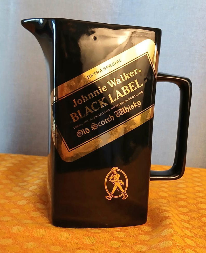 May include: Black ceramic pitcher with a handle and spout. The pitcher features a gold and black label with the text "Johnnie Walker BLACK LABEL Old Scotch Whisky". A gold emblem of a man walking is also visible.