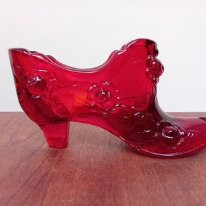 May include: A red glass shoe with a high heel and floral design. The shoe is made of clear glass with a red tint and has a floral pattern etched into the surface.