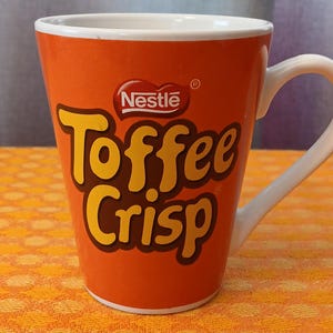 May include: An orange ceramic mug with the Nestle logo and the words "Toffee Crisp" in yellow and brown. The mug has a white handle and a white interior. The mug is sitting on an orange patterned surface.