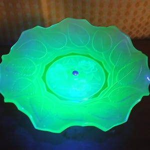 May include: A round, translucent green glass bowl that glows under ultraviolet light. The bowl features a scalloped edge and a decorative relief pattern of leaves around the rim, with a central circular design depicting floral motifs.