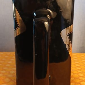 May include: Black rectangular mug with a glossy finish and a curved handle. Gold and white accents are visible on the sides. The word "DELUXE" is printed on the side of the mug.