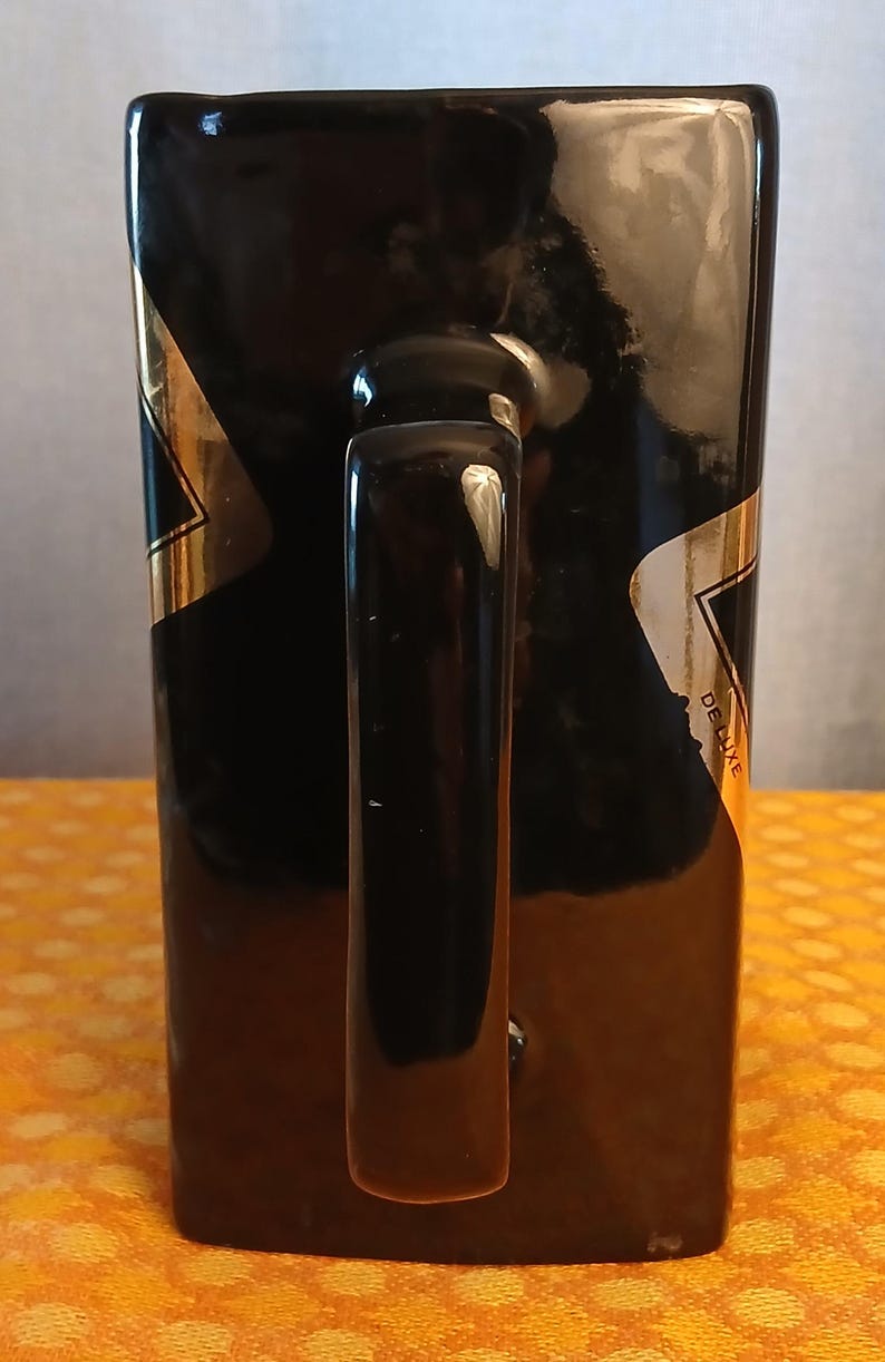 May include: Black rectangular mug with a glossy finish and a curved handle. Gold and white accents are visible on the sides. The word "DELUXE" is printed on the side of the mug.