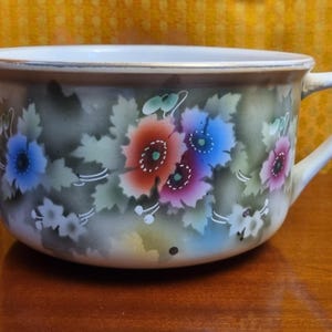May include: A white ceramic mug with a floral design. The flowers are painted in shades of blue, red, and pink. The mug has a wide, rounded shape and a handle.