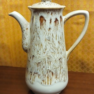 May include: A tall, ceramic coffee pot with a lid and handle. The pot is primarily white with brown drip glaze patterns. The handle is curved, and the spout is slender. The lid has a small knob on top.