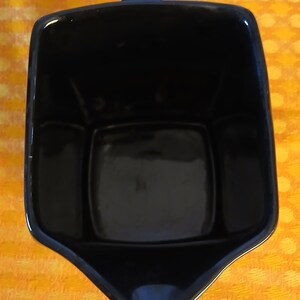 May include: A black ceramic creamer with a handle. The creamer has a square shape with a spout and a handle on the top. The creamer is sitting on a yellow and orange patterned surface.