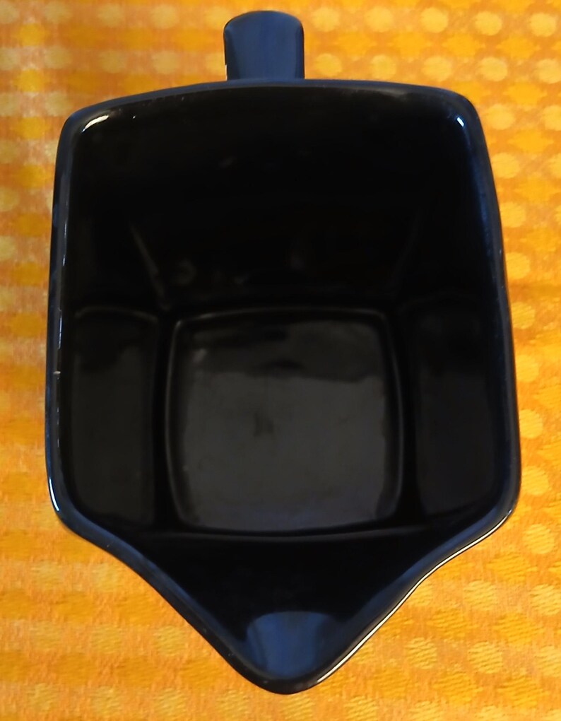 May include: A black ceramic creamer with a handle. The creamer has a square shape with a spout and a handle on the top. The creamer is sitting on a yellow and orange patterned surface.