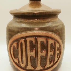 May include: A ceramic coffee canister with a lid. The jar is a mottled brown color with a lighter brown oval that reads "COFFEE" in raised letters. The lid has a small knob.