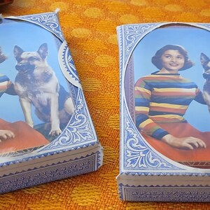 May include: Two vintage playing card boxes. Each box features a portrait of a woman in a striped sweater and red skirt, with a German Shepherd dog. The boxes have a blue and white decorative border.