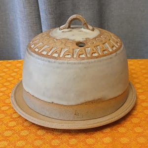 May include: A ceramic cheese dome with a matching plate. The dome is off-white with a tan base and a decorative pattern around the top. It has a small handle and a hole for ventilation. The plate is also tan.