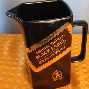 May include: Black ceramic pitcher with a handle, featuring a gold and black label that reads "Johnnie Walker BLACK LABEL Old Scotch Whisky." The label includes a graphic of a man walking. The pitcher has a unique angled design.
