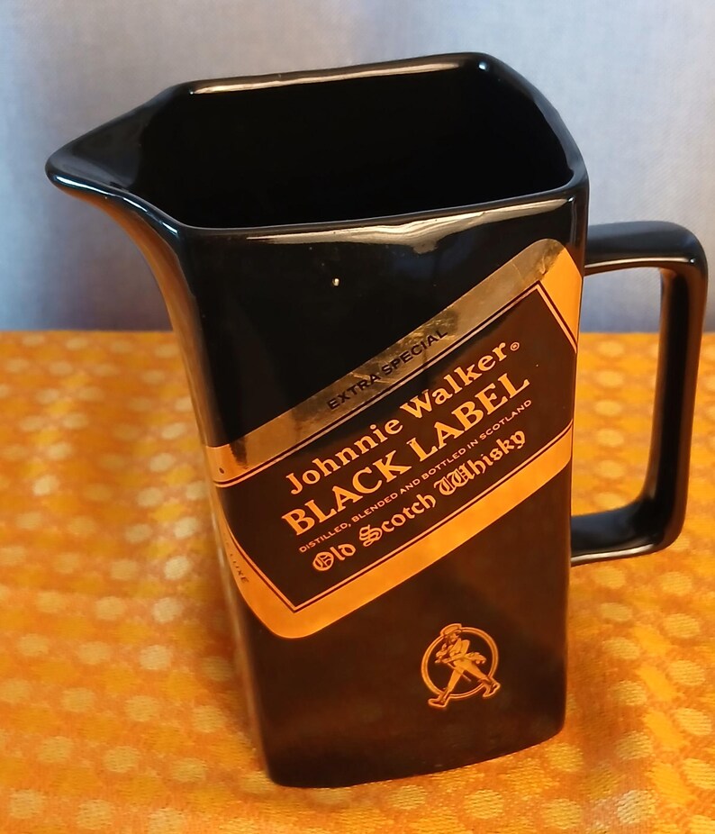 May include: Black ceramic pitcher with a handle, featuring a gold and black label that reads "Johnnie Walker BLACK LABEL Old Scotch Whisky." The label includes a graphic of a man walking. The pitcher has a unique angled design.
