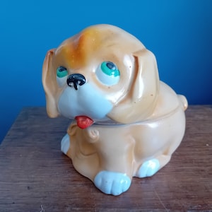May include: A ceramic dog figurine with a brown and white body, green eyes, and a red tongue sticking out. The figurine is sitting on a wooden surface.