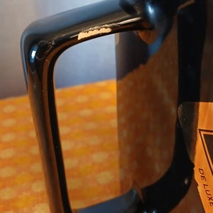 May include: Close-up of a black mug with a square handle. The handle has a glossy finish with a small area of chipped paint. A partial view of a label with the words "DE LUXE" is visible on the side of the mug.