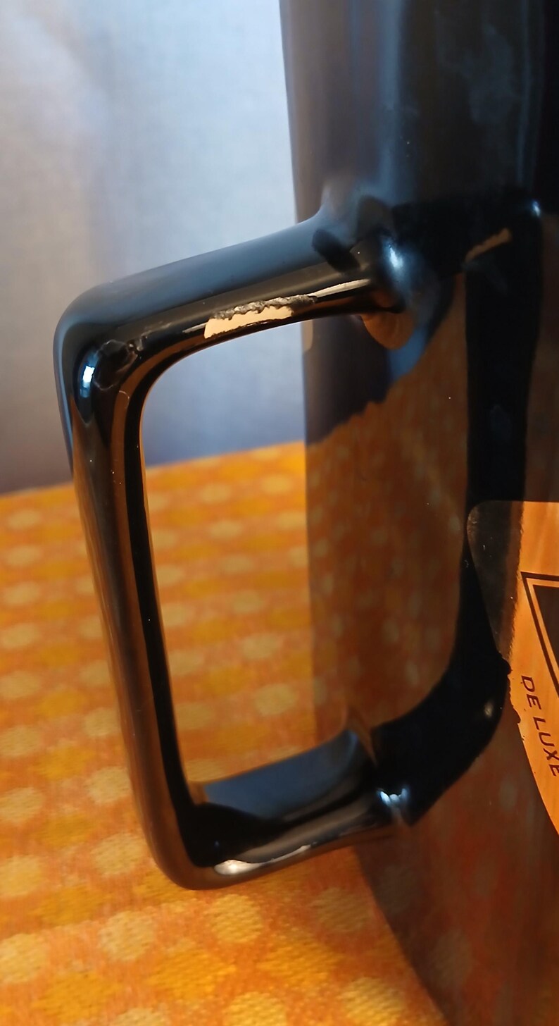 May include: Close-up of a black mug with a square handle. The handle has a glossy finish with a small area of chipped paint. A partial view of a label with the words "DE LUXE" is visible on the side of the mug.