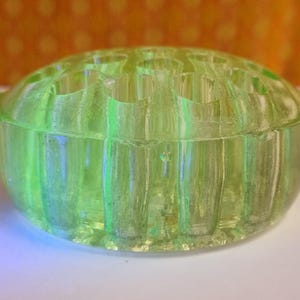 May include: A round, light green glass flower frog with a ribbed exterior and multiple holes on top. The vintage floral arranging tool is made of translucent glass and sits on a white surface.