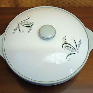 May include: A round, white ceramic casserole dish with a matching lid. The lid features a central light blue knob and two symmetrical, stylized green and black leaf-like patterns. The dish has integrated side handles and a light blue rim.