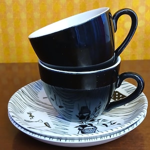 May include: Two stacked ceramic cups and two matching saucers. The top cup is solid black with a white interior. The bottom cup and saucers feature a white base with black line drawings of various household objects and plants.