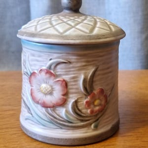 May include: A ceramic jar with a lid, decorated with a floral motif. The cylindrical jar has a textured, pale body, embellished with embossed pink and red flowers. The lid features a diamond pattern and a small knob.