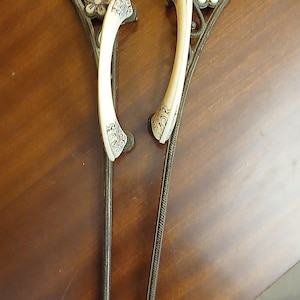 May include: Two matching metal drawer pulls featuring an ornate, curved bronze-toned frame with a floral motif at the top and a smooth, cream-colored arched handle insert. Each pull has a long, slender metal rod extending downward from the base.