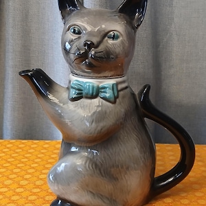 May include: A gray ceramic teapot shaped like a cat with a black tail and handle. The cat has a white collar and a teal bow tie. The teapot has a spout and a lid that is the cat's head.