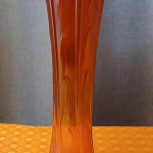 May include: An orange iridescent glass vase with a flared rim and a rounded base. The vase has a tall, slender shape with vertical ridges and a decorative pattern. The vase is approximately 25 cm tall.