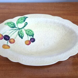 May include: Oval, off-white ceramic serving dish with a scalloped edge. It features a sprig of green leaves and cherries in yellow, orange, and purple. The dish has a crackle glaze. Measurements are in centimetres.