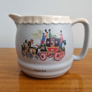 May include: A white ceramic pitcher with a cream-colored rim and a handle. The pitcher features a colorful illustration of a horse-drawn carriage with passengers and the text "Lands End".
