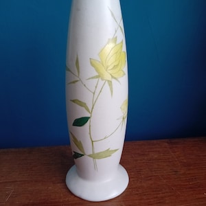 May include: A white ceramic vase with a yellow rose and green leaves design.