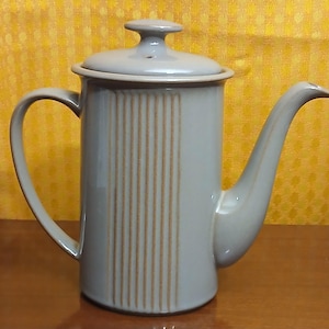 May include: A light gray ceramic coffee pot with a lid and a long spout. Vertical brown stripes decorate the front of the pot. The handle is curved, and the pot sits on a wooden surface.