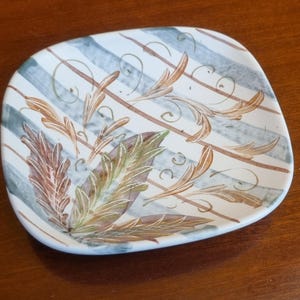 May include: A square ceramic plate with a white base, featuring blue and brown stripes and decorative leaf patterns in brown, green, and purple. The plate has rounded corners and is set on a wooden surface.