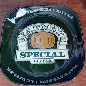 May include: A green ceramic ashtray with a white border and the words "Watneys Special Bitter" printed around the edge. The center of the ashtray features a stylized image of a barrel with the words "Watneys Special Bitter" printed below it.