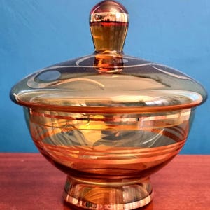 May include: A vintage amber glass candy dish with a lid. The dish features horizontal gold stripes and a decorative gold finial on the lid. The glass has a reflective quality, and the dish sits on a pedestal base.
