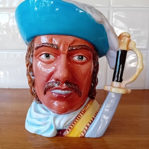 May include: A ceramic mug shaped like a man's head wearing a blue and white hat. The mug has a sword-shaped handle.