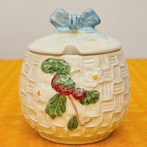 May include: A cream-coloured ceramic jar with a lid, featuring a woven basket design. The jar is adorned with a strawberry and floral motif, and a light blue bow on the lid. The jar is designed for kitchen use.