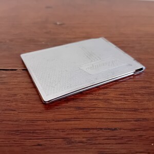 May include: A silver metal compact with a textured surface and the word "Germany" engraved on the front.